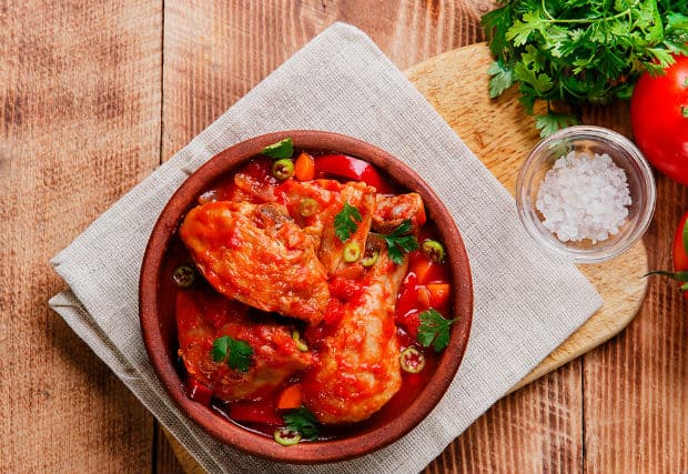 курица, тушенная с овощами, в глиняной посуде на доске, застеленной салфеткой, рядом пучок зелени и соль