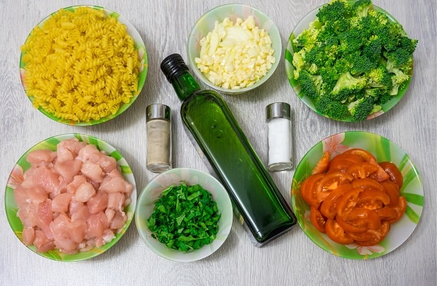 нарезанные помидоры, брокколи, зелень, куриное филе, лук и чеснок в тарелках, рядом макароны на тарелке, солонки и бутылка масла