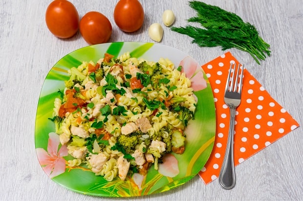 паста с курицей, овощами и зеленью на тарелке, рядом вилка на салфетке в горошек, помидоры, зубчики чеснока и зелень