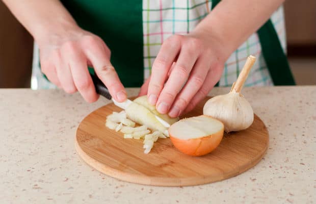 репчатый лук нарезается ножом, рядом половинка луковицы и головка чеснока на разделочной доске на столе