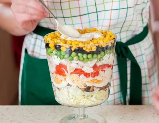 слоеный салат из шампиньонов, курицы, яиц, консервированного горошка и кукурузы в салатнице на ножке на столе